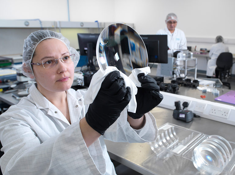 woman wearing glassing in white coat holding up and inspecting large optical glass