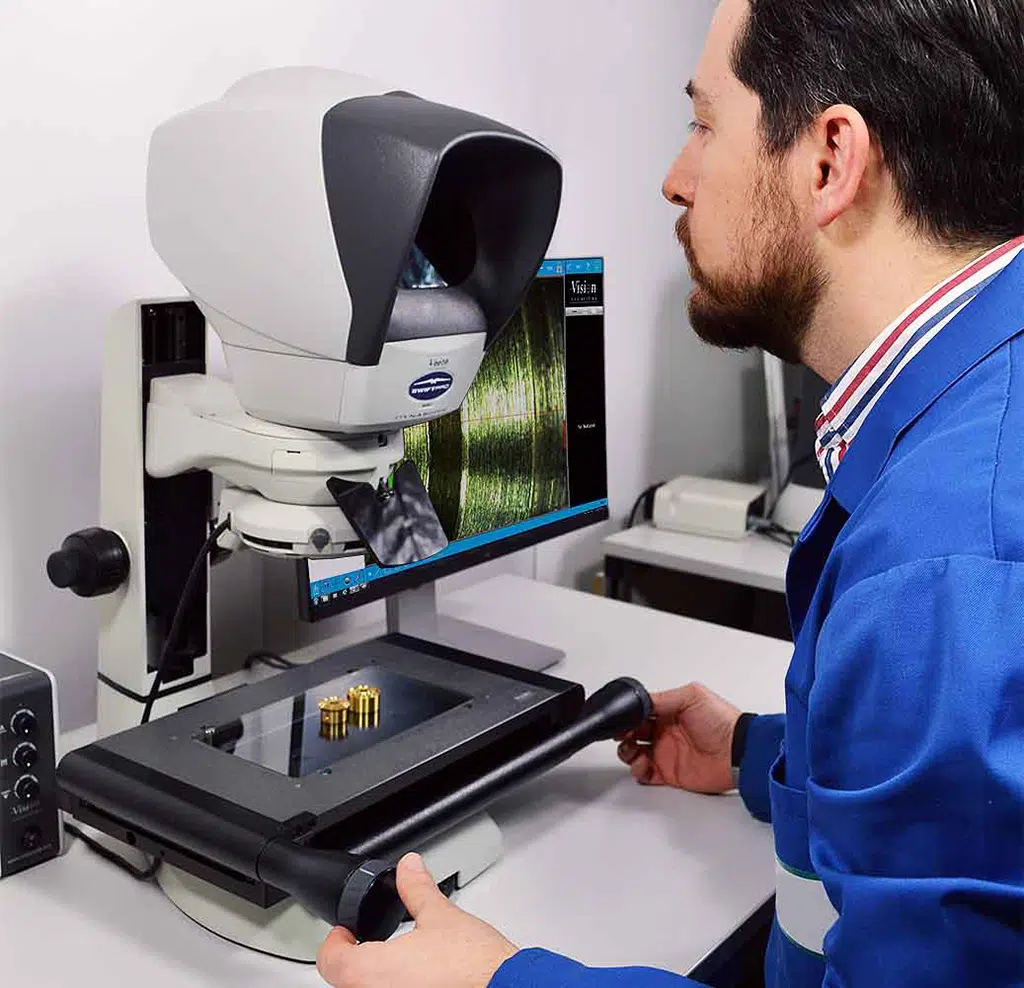 Operator viewing small brass components through an eyepiece-less optical head, with a magnified image displayed on an adjacent monitor