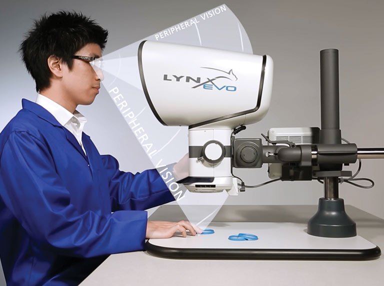 Operator sitting upright at a Lynx EVO eyepiece-less stereo microscope, inspecting small parts with peripheral vision maintained