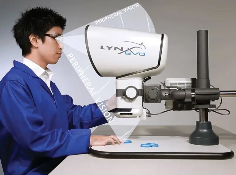 Operator sitting upright at a Lynx EVO eyepiece-less stereo microscope, inspecting small parts with peripheral vision maintained